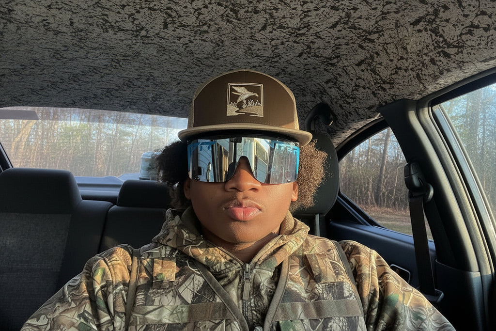 Person wearing a cap and sunglasses inside a vehicle with a camouflage-patterned ceiling.